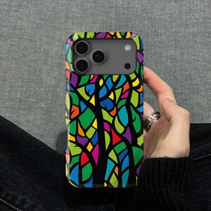 May include: A phone case with a stained-glass tree design. The case has a black background with a vibrant tree composed of green, blue, yellow, orange, and purple shapes. The phone case is held in a person's hand.