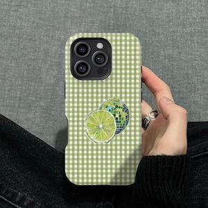 May include: A phone case with a green and white gingham pattern. The case features a lime slice and a disco ball design. The phone case is held by a person.
