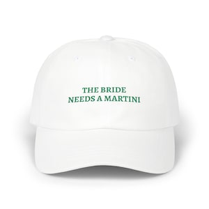 May include: White baseball cap with a curved brim. The cap features the text "THE BRIDE NEEDS A MARTINI" in green block letters across the front. The hat is a classic, casual style.