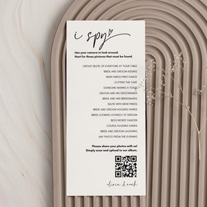 May include: A white card with the text "i spy" written in black cursive. The card lists a series of things to find in a wedding photo, such as "bride and groom first dance" and "someone cutting the cake". A QR code is at the bottom of the card.