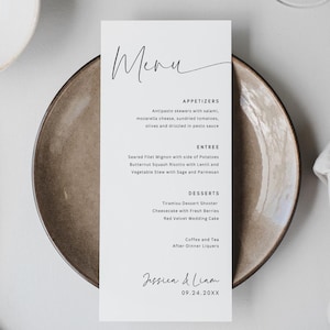 May include: A minimalist wedding menu card with the word "Menu" in elegant script. The menu lists appetizers, entrees, and desserts. A gold fork and knife are next to a gray napkin. A white plate and a decorative leaf are also visible.