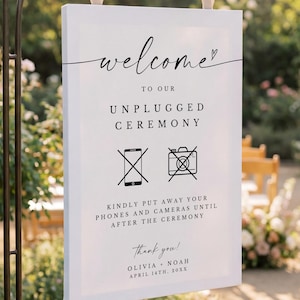 Puede incluir: Un letrero blanco con las palabras "welcome" y "Unplugged Ceremony" en escritura negra. El letrero también incluye símbolos de teléfonos y cámaras con una línea que los atraviesa, y los nombres Olivia y Noah.