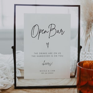 May include: A black framed sign with a white background that reads "Open Bar" with a heart symbol. The text below reads "The drinks are on us, the hangover is on you." The sign also includes the text "Cheers!" and the names "Nicole & Liam" with the date "09.24.20XX".