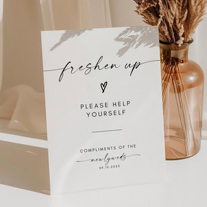以下が含まれることがあります： 「freshen up」という黒い文字が書かれた白い看板。ハートマークと「どうぞご自由にお使いください」の文字も書かれている。文字の下には「Compliments of the newlyweds 08.10.2023」と書いてある。