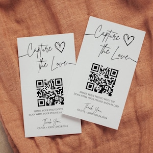 May include: Two white cards with the text "Capture the Love" and a heart symbol. Each card has a QR code and the text "Share your photo with us! Scan with your phone and upload." The cards also include the text "Thank You, Olivia + John 09.24.2024."