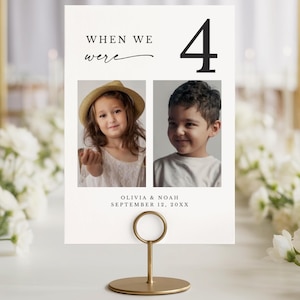 May include: A table number card with the text "When We Were" and the number 4. The card features two photos of children and the names Olivia & Noah with the date September 12, 20XX. The card is held by a gold metal stand.