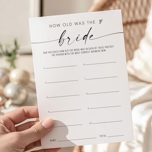 May include: A white card with the text "HOW OLD WAS THE bride" in black script. The card includes lines for answers and prompts, "CAN YOU GUESS HOW OLD THE BRIDE WAS IN EACH OF THESE PHOTOS? THE PERSON WITH THE MOST CORRECT ANSWERS WIN!"