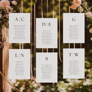May include: A gold metal wedding seating chart with white cards suspended within a circular frame. The cards are adorned with names and table assignments, surrounded by floral arrangements of pink and white flowers.