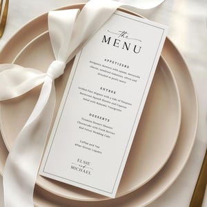May include: A wedding menu card with a white ribbon tied around a stack of plates. The menu features a minimalist design with the text "The Menu" at the top and a list of appetizers, entrees, and desserts. Gold cutlery is visible.