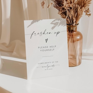 May include: A white rectangular sign with the words "freshen up" in cursive, a heart symbol, and the text "PLEASE HELP YOURSELF." The sign also includes "COMPLIMENTS OF THE newlyweds" and the date 09.10.2025. A decorative vase with dried flowers is in the background.