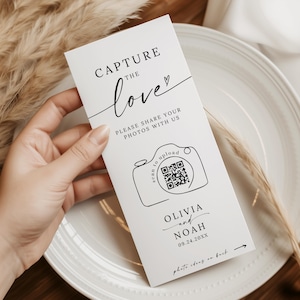 May include: A white wedding card held by a person. The card features the text "CAPTURE the love" in a script font, along with "PLEASE SHARE YOUR PHOTOS WITH US." A camera graphic with a QR code is also present, along with names and a date.