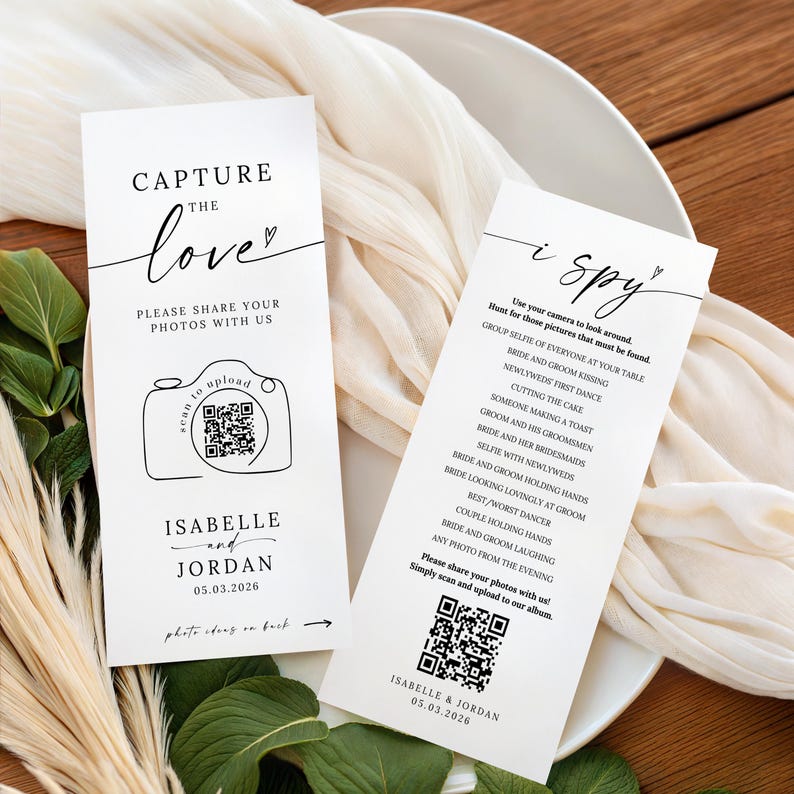 May include: Two white wedding cards with black text. One card reads "Capture the love" with a camera graphic and QR code. The other card says "I spy" with a list of photo prompts. Both cards include the names "Isabelle and Jordan" and the date 05.03.2026.