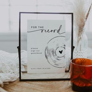 May include: A framed sign with the text "FOR THE record" and "please sign our guestbook". The sign features a black and white vinyl record graphic with a heart. The frame is black and the sign is on a wooden surface.