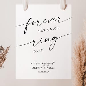 Pode incluir: Uma placa branca com a frase "forever has a nice ring to it" em elegante escrita preta. A placa também inclui o texto "we're engaged!" com nomes e uma data, perfeita para uma celebração de casamento ou noivado.