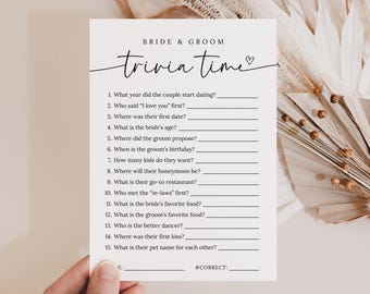 May include: A white card with the text "BRIDE & GROOM trivia time" in elegant script. The card features fill-in-the-blank questions about the couple, perfect for a wedding game. The card is held by a hand, with dried flowers in the background.