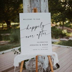May include: A white wedding sign on a wooden easel. The sign reads "Welcome to our happily ever after" in elegant script, celebrating Jessica & Noah in Austin, TX, on 09.20.20XX. A white cloth drapes the bottom of the sign.