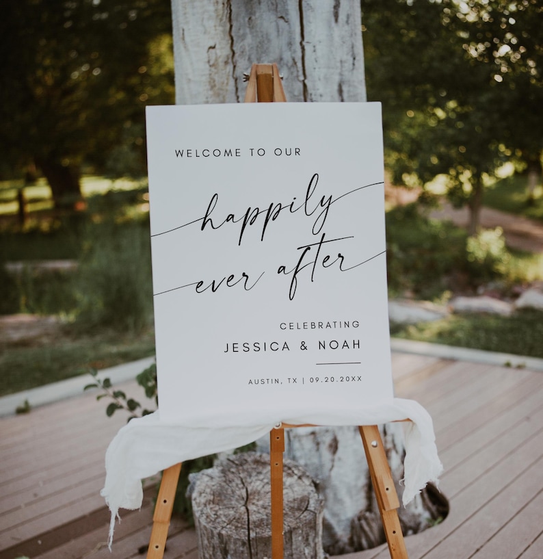 May include: A white wedding sign on a wooden easel. The sign reads "Welcome to our happily ever after" in elegant script, celebrating Jessica & Noah in Austin, TX, on 09.20.20XX. A white cloth drapes the bottom of the sign.