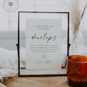 May include: A framed sign with the text "CAN'T WAIT TO SEE WHAT develops" in a stylish font. The sign includes instructions for capturing special moments. A decorative glass and a vase with dried flowers are in the background.