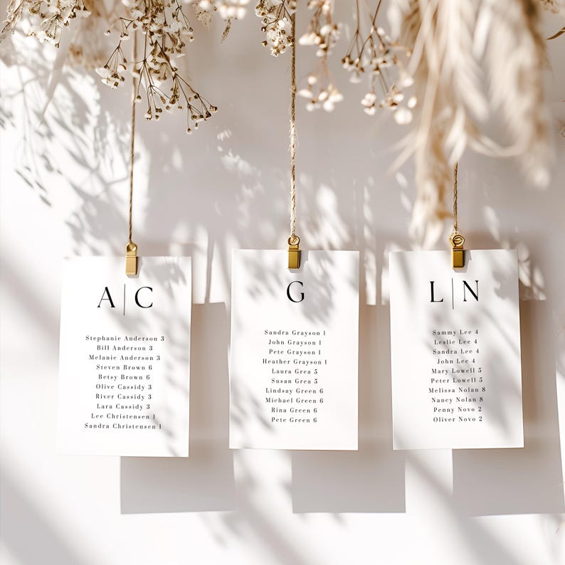 May include: Elegant wedding seating chart featuring three white cards suspended by gold clips and twine. Each card displays guest names and table numbers in a minimalist font. The cards are adorned with floral accents, creating a sophisticated aesthetic.