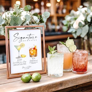 May include: A rose gold framed sign titled Signature Cocktails with watercolor illustrations of a lemon drop and an old fashioned. The sign lists ingredients for Hers and His drinks, plus the names Olivia and Noah and the date 9.20.2026.