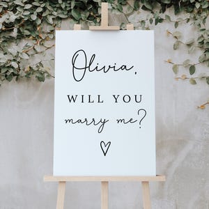 Puede incluir: Un letrero blanco sobre un caballete de madera con las palabras "Olivia, WILL YOU marry me?" en escritura negra y un símbolo de corazón. El fondo presenta follaje verde y una pared de color claro.