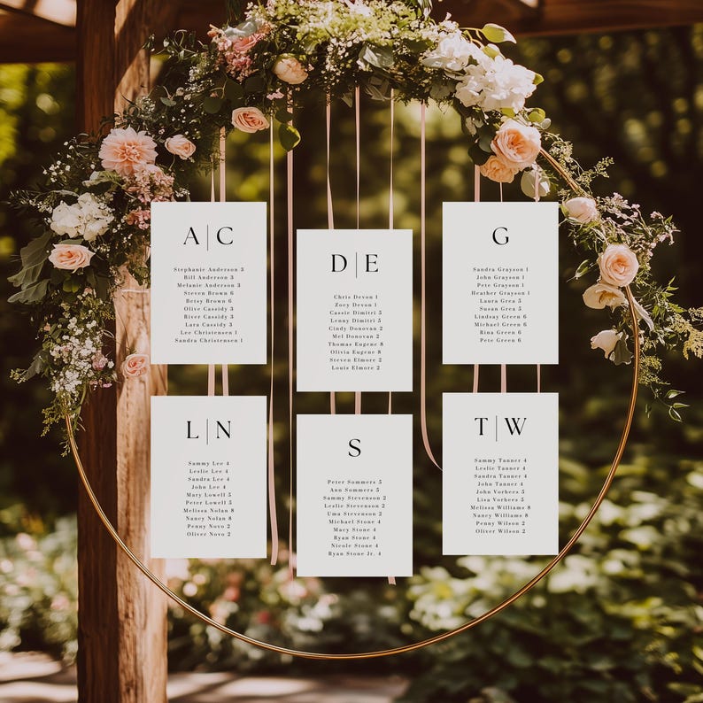 May include: A gold metal wedding seating chart with white cards suspended within a circular frame. The cards are adorned with names and table assignments, surrounded by floral arrangements of pink and white flowers.