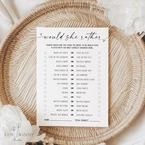 Peut inclure: Une carte blanche avec le texte "would she rather" et une liste de choix pour un jeu de douche nuptiale. La carte est sur un plateau tissé avec une fleur blanche dans le coin. Le jeu comprend des options comme "Se lever tôt ou rester tard."