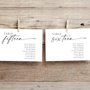 May include: Two white cards with black text, clipped to a string with wooden pegs. The cards read "TABLE fifteen" and "TABLE sixteen" in elegant script. Below the numbers are lists of names. The background is a rustic wooden surface.
