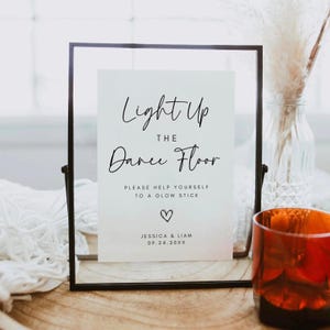 Op de afbeelding: Een rechthoekig zwart metalen frame met een wit bordje met zwarte tekst: Light Up THE Dance Floor PLEASE HELP YOURSELF TO A GLOW STICK, een harticoontje en de namen JESSICA & LIAM met de datum 09.24.20XX eronder gedrukt.