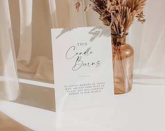May include: A white card with black text that reads "This Candle Burns" and "In loving memory of those who are not here with us today, but will forever remain in our hearts". The card is sitting on a white surface with a vase of dried flowers in the background.