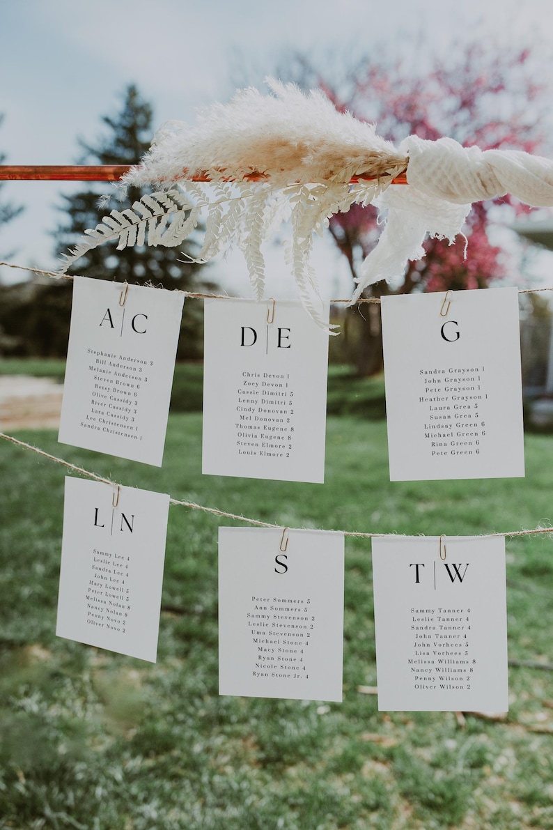 May include: Wedding seating chart with white cards clipped to a string. Each card has a letter and guest names with table numbers. The cards are decorated with pampas grass and greenery.