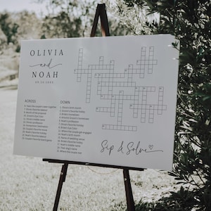 May include: A white sign on a wooden easel featuring a crossword puzzle. The sign includes the names "Olivia and Noah" with a wedding date. The words "Across" and "Down" are listed with clues. The phrase "Sip & Solve" is written at the bottom.