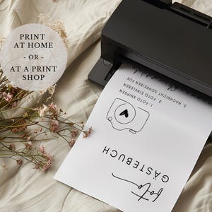 May include: A white sheet of paper with black text and a simple camera illustration. The text reads "G&Auml;STEBUCH", "1. FOTO KNIPSEN", "2. FOTO EINKLEBEN", "3. NACHRICHT SCHREIBEN F&Uuml;R". The paper is being fed into a black printer.