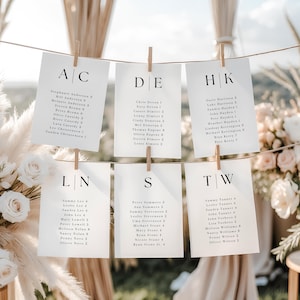 May include: White cardstock seating chart with names and table assignments, clipped to a string with wooden clothespins. The cards are arranged in rows, with each card featuring a letter combination at the top.