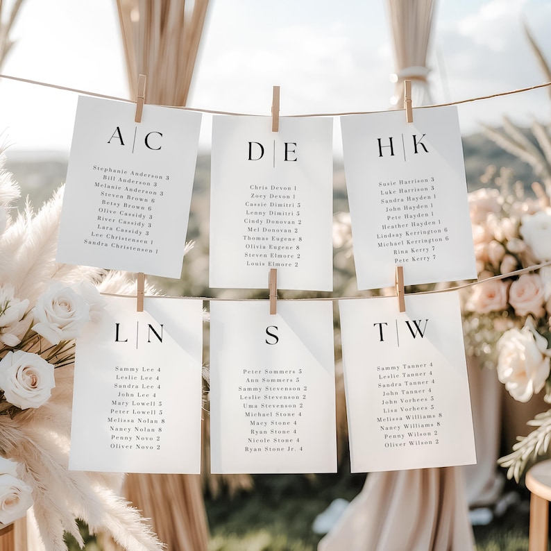 May include: White cardstock seating chart with names and table assignments, clipped to a string with wooden clothespins. The cards are arranged in rows, with each card featuring a letter combination at the top.