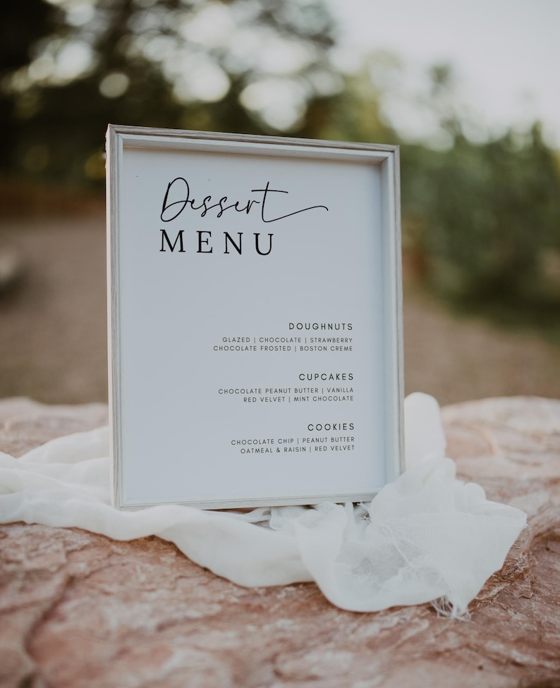 May include: A white sign with black text listing a dessert menu. The menu includes doughnuts, cupcakes, and cookies with different flavors. The sign is sitting on a rock with a white cloth draped over it.