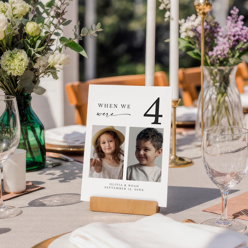 May include: A white table number card with the text "WHEN WE were" and the number "4" in black. Two small photos of children are below the text. The card is held in a wooden block. The text "OLIVIA & NOAH SEPTEMBER 12, 20XX" is at the bottom.
