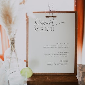 May include: A white sign with a brown wooden frame and a clip at the top. The sign says "Dessert Menu" in black cursive writing. The menu lists different types of desserts, including doughnuts, cupcakes, and cookies.