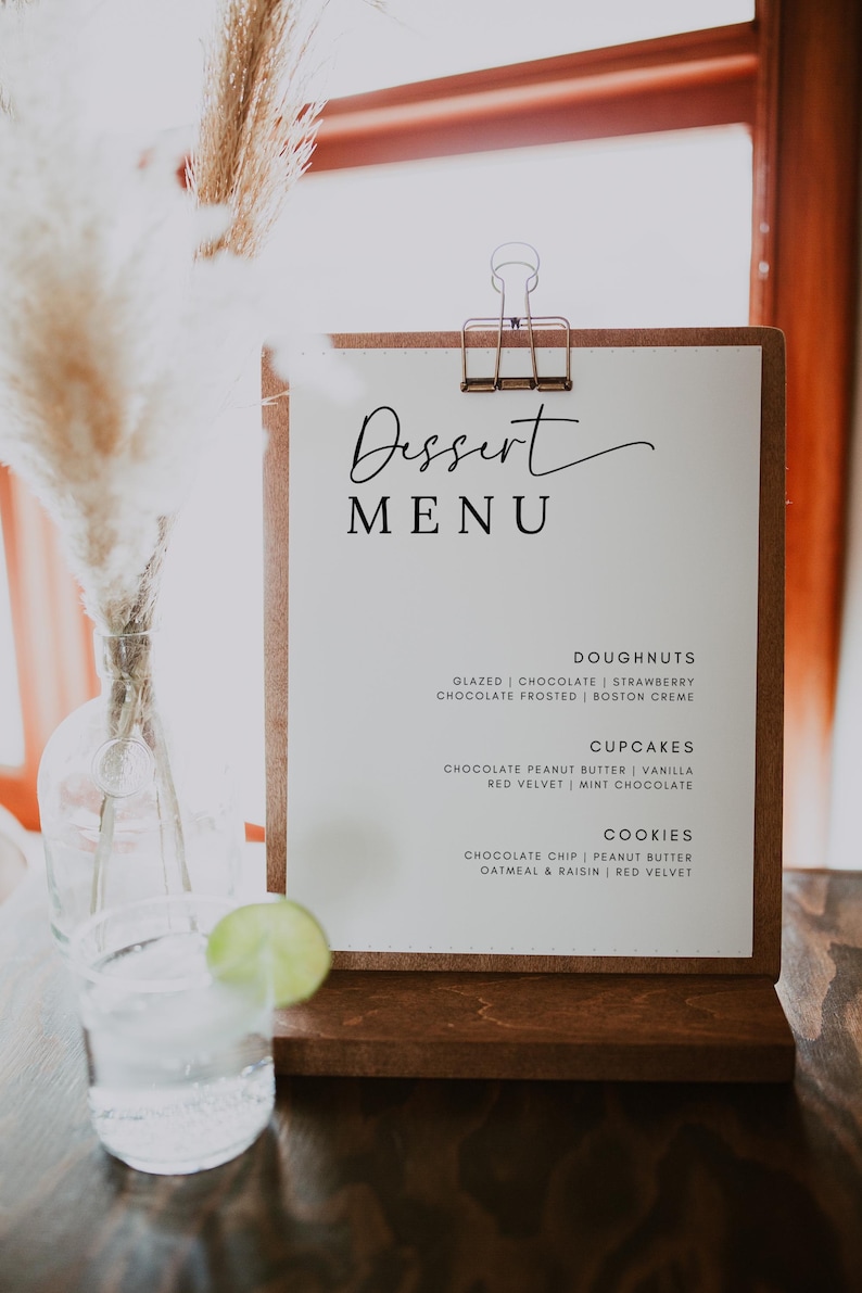 May include: A white sign with a brown wooden frame and a clip at the top. The sign says "Dessert Menu" in black cursive writing. The menu lists different types of desserts, including doughnuts, cupcakes, and cookies.