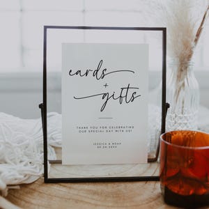 May include: A framed sign with the words "cards + gifts" in elegant script. The sign also includes the text "THANK YOU FOR CELEBRATING OUR SPECIAL DAY WITH US!" and the names Jessica & Noah with a date. A decorative wedding sign.