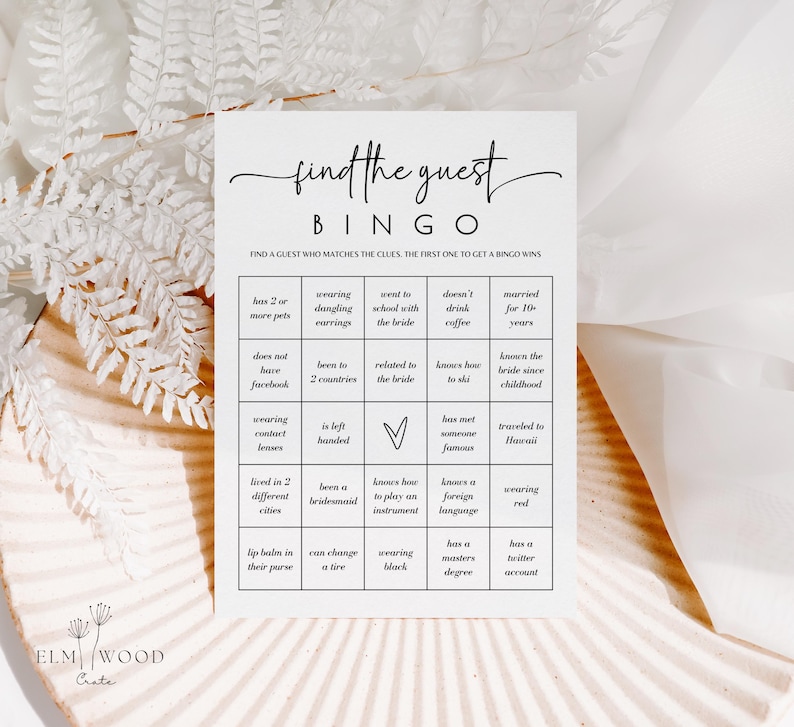 May include: A white Bingo game card with the text "find the guest" in elegant script. The card features a grid with various prompts like "wearing dangling earrings" and "knows how to ski". The card is designed for a wedding or bridal shower game.