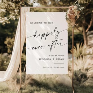 May include: A white wedding sign with the words "Welcome to Our Happily Ever After" in elegant black script. The sign also includes the names Jessica & Noah, and the location and date. The sign is on a gold metal stand, decorated with flowers.