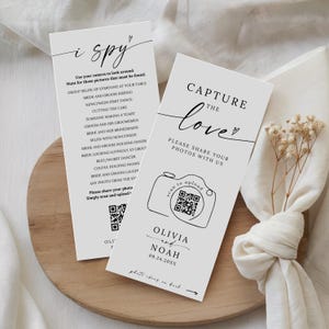 May include: Two white wedding signs with black text. One sign says "i spy" with photo prompts. The other says "Capture the love" with a QR code to upload photos. Names and a date are printed below the camera graphic.