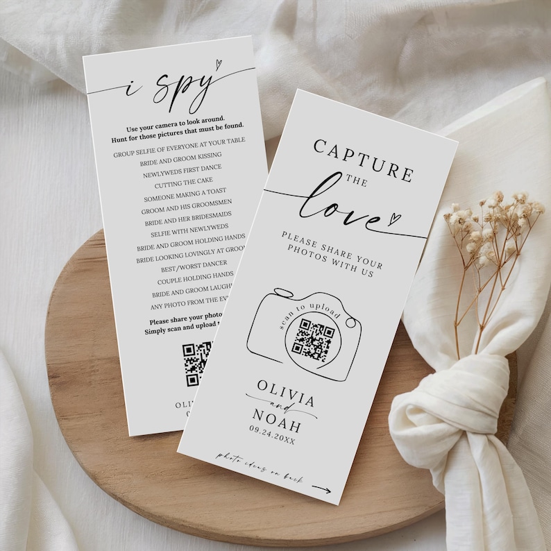 May include: Two white wedding signs with black text. One sign says "i spy" with photo prompts. The other says "Capture the love" with a QR code to upload photos. Names and a date are printed below the camera graphic.