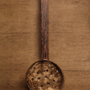Può includere: Utensile da cucina in legno con una ciotola rotonda e perforata e un lungo manico. Il manico è marrone scuro con una venatura strutturata e la ciotola è marrone chiaro con numerosi piccoli fori. Per filtrare o raccogliere.
