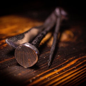 May include: Close-up of three metal tools on a wooden surface. The tools include a nail with a rounded head and two other tools with sharp edges. The wood has a warm, orange-brown colour with visible grain.