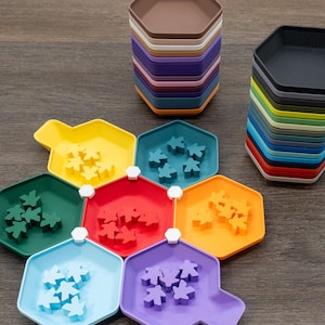 May include: A collection of hexagonal plastic trays in various colors, including yellow, teal, green, red, orange, light blue, and purple. Each tray contains matching colored wooden meeple game pieces. Two tall stacks of empty hexagonal trays are also visible.