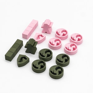 May include: A collection of game pieces in light pink and olive green. The set includes two human-shaped meeples, two rectangular bars made of four connected cubes, two shield-shaped tokens with horse silhouettes, and eight circular tokens with dragon cutouts.