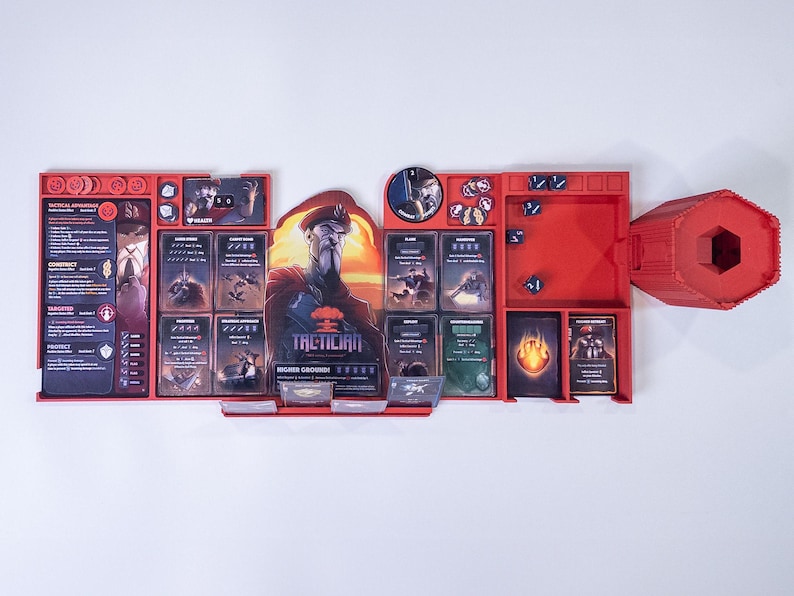 May include: A red plastic board game organizer featuring a central character illustration labeled Tactician, multiple slots for game cards, small circular tokens, and six-sided dice. The organizer includes a separate red hexagonal dice tower.
