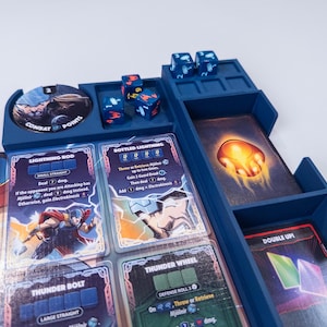 May include: A blue plastic board game organizer containing four six-sided dice with colored symbols, a circular combat points token, and several rectangular game cards featuring illustrations of a character with a hammer and lightning effects.